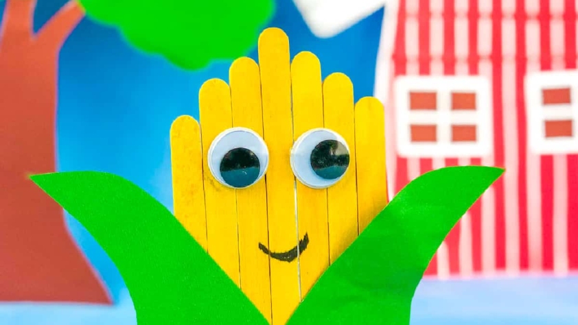 craft sticks painted yellow and glued side by side to resemble a corn cob with googly eyes. Green construction paper is cut to look like leaves and glued on the cob
