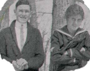 black and white photo; boy smiling with hands clasped as if holding something, and girl smiling with arms crossed